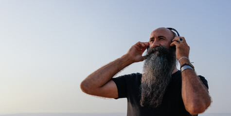 Portrait of a virile man with a long espectacular beard wearing black t-shirt listening music with his hands on his head at ear level in a blue clear sky background. © anuatmoralesfoto