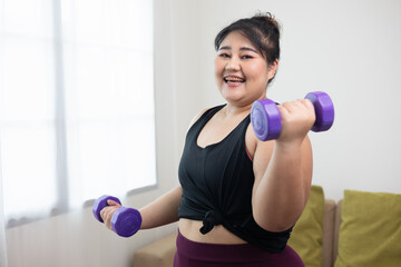 Fototapeta na wymiar Attractive asian young fitness woman plus size lifting dumbbell weights workout at home in living room. Female training and exercise wearing sport wear fit body. Happy and is proud of herself.