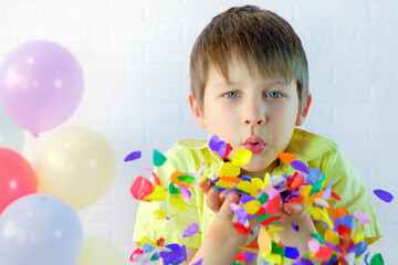Fototapeta na wymiar the boy blows multi-colored confetti from his hands. A child in a yellow shirt against a white wall and balloons. Portrait of a caucasian boy 7-8 years old. Concept - Happy birthday, party, holiday