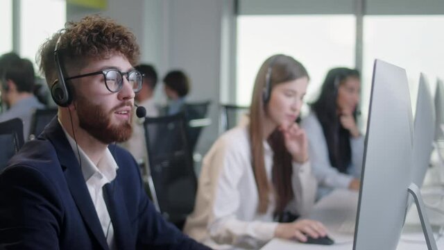 Call Center Office: Portrait of Friendly Caucasian Male Technical Customer Support Specialist Talking on a Headset, Diverse group of workers talks on sale hotline. Busy seller at agency office indoor