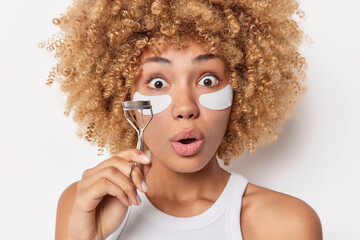 Amazed astonished curly haired young woman applies patches under eyes stares bugged eyes keeps mouth opened uses eyelasher curler undergoes beauty procedures isolated over white studio background.