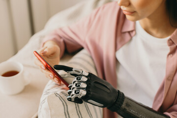 Crop shot of woman holding mobile phone of red color in bionic prosthesis hand leaned over sofa...