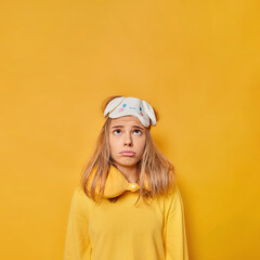 Vertical shot of frustrated sleepy woman looks above unhappily wears sleepmask neck pillow and casual jumper cannot fall asleep has sleep disorder isolated over yellow background copy space.