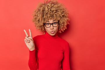 Romantic curly haired young woman keeps lips rounded makes peace gesture or victory sign wears transparnt spectacles and casual turtleneck isolated over red background. Body language concept