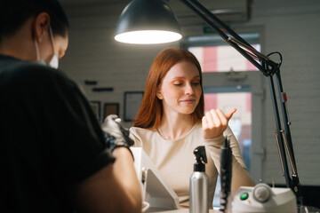 Smiling young woman examining treated nails and assessing work of manicurist in cosmetology salon. Professional female cosmetologist using file to make beautiful oval shape of nails doing manicure.