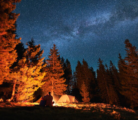 Rear view of couple in love relaxing in their camp at evening. Vacationers sitting by campfire and looking at the starry sky with Milky way. Romantic atmosphere during camping at starry night outdoors