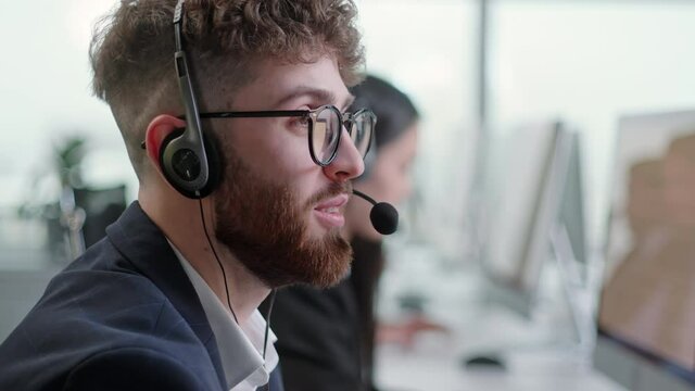 Close Up Portrait Man of a Technical Customer Support Specialist Talking on a Headset while Working on a Computer in a Call Center. Diverse group of workers talks on sale hotline. Busy seller at