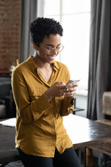 Happy African millennial professional girl in glasses reading text message on smartphone, smiling,...