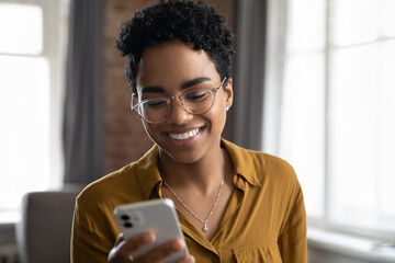Happy Afro American young business woman in glasses using smartphone, chatting online with toothy smile, reading sms with good news, typing text message, using virtual ecommerce app for shopping - obrazy, fototapety, plakaty