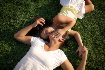 Una niña besando a su madre tumbada en la hierva al aire libre sin mascarilla sonriendo tomando el sol con ropa clara y cabello moreno.
