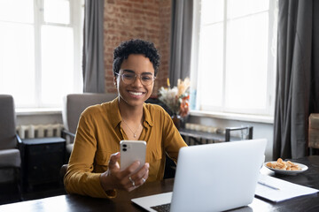 Happy African business woman chatting on digital gadgets online, holding mobile phone, using laptop computer, looking at camera, smiling. Freelance employee at home workplace head shot