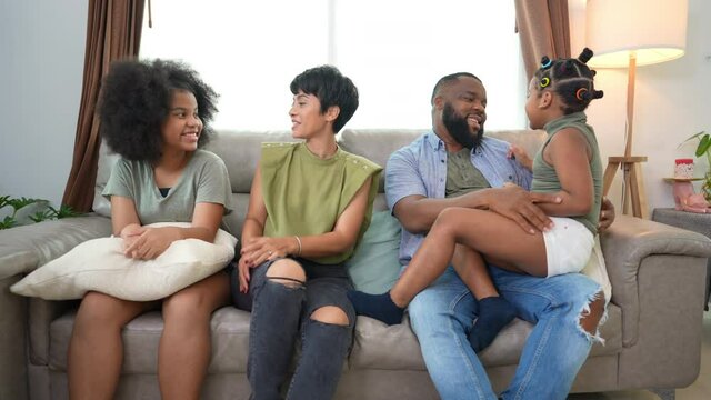 Happy African family parents and two little daughter sitting on sofa in living room playing and talking together. Father and mother with child girl kid enjoy indoor lifestyle activity together at home