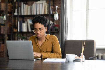 Focused millennial Afro American business woman working at laptop at home office table. Young...