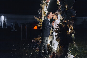 A stylish groom and a beautiful bride in a long dress stand holding hands, imitating a mirror, at night near a luminous reed arch decorated with lamps and garlands.