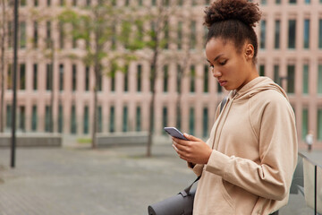 Outdoor shot of serious woman with combed hair dressed in casual hoodie uses mobile phone chats online carries rolled karemat poses against blurred building has self training. People sporty lifestyle