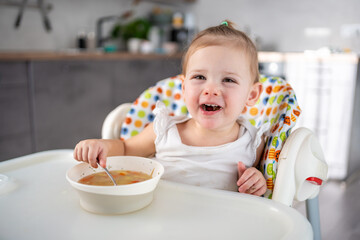 Cute baby girl toddler sitting in the high chair and eating her lunch soup at home kitchen.