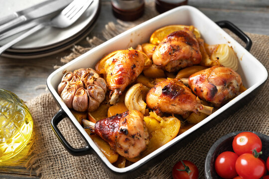Baked chicken legs and potatoes with oranges, garlic and onions in a baking dish on a light gray wooden culinary background closeup