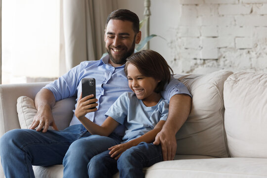 Happy son kid and cheerful laughing dad talking on video call, watching media content on internet, making selfie for social networks, using online app for playing video games, shopping