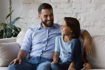Cheerful dad talking to happy adopted son kid, sitting on couch with boy, chatting, laughing,...