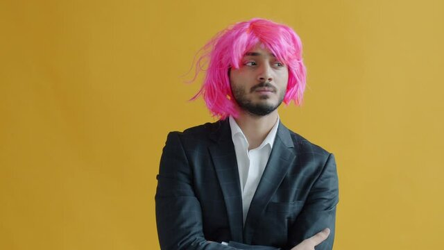 Slow motion portrait of mixed race man taking off pink wig and leaving feeling unhappy on yellow color background. Boring party and negativity concept.
