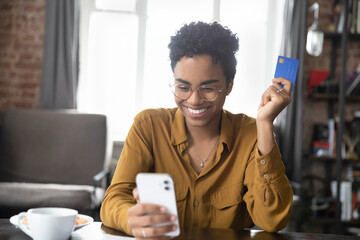 Happy cheerful millennial Black girl enjoying online shopping, using ecommerce app on smartphone, paying for purchase, order by credit card, buying goods on Internet. Finance, spending money concept