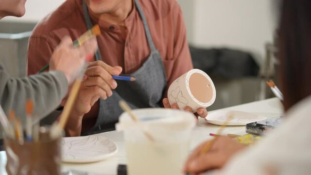 Asian Lgbtq guy design stylish pattern for painting self-made pottery mug with friends. Happy man enjoy leisure activity handicraft hobbies ceramic sculpture painting workshop at pottery studio