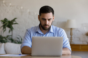 Focused millennial business man using laptop at table. Young entrepreneur, freelancer in casual,...