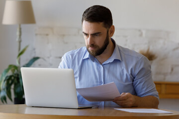 Fototapeta premium Focused millennial business man checking paper reports. Entrepreneur reviewing financial document at laptop. Homeowners paying rent bills, invoice, reading mortgage, loan, credit notice from bank,