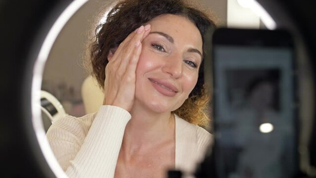 Beautiful middle aged woman demonstrates the application of a moisturizing and nourishing cosmetic product for skin care. Beauty blogger present beauty cosmetics sitting in front camera for recording