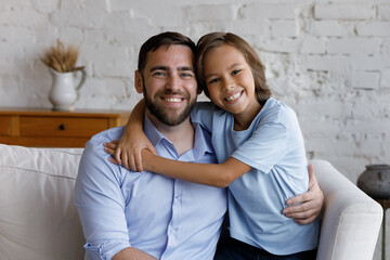 Happy loving father and cheerful adopted preteen son hugging on couch with love, affection, tenderness, looking at camera with toothy smiles, hugging, enjoying friendship, family leisure