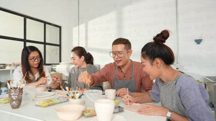 Group of Happy Asian man and woman friends enjoy and having fun indoor lifestyle painting self-made pottery together at pottery studio. Hobby leisure activity handmade ceramic art painting workshop.