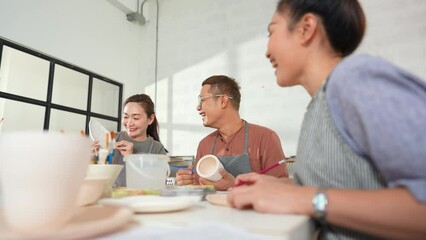 Group of Happy Asian man and woman friends enjoy and having fun indoor lifestyle painting self-made pottery together at pottery studio. Hobby leisure activity handmade ceramic art painting workshop.