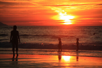 Madre y niños en la Playa jugando en la orilla al anochecer con cielo anaranjado.