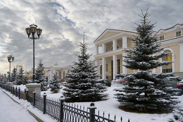 One of the city alleys with lanterns, a low forged fence and snow-covered firs in Yekaterinburg (Russia) on a cloudy winter day. Old restored buildings of white and yellow color. Cityscape with snow 