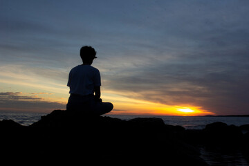 Girl sitting on a rock meditating in the late afternoon near the sea, sun setting in the background. Unrecognizable silhouette. Amazing landscape at dusk. Stones, ocean and sky. Meditation.