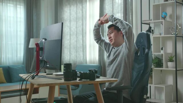 Asian Cameraman In Long Sleeved T-Shirt And Black Pants Stretching And Leaning Back While Using Desktop Computer For Working At Home.
