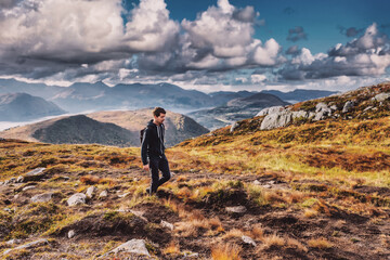 Young adult man hiking on top of a mountain in norway during autumn, wearing a backpack, looking down, sky and horizon with mountain range, volda norway