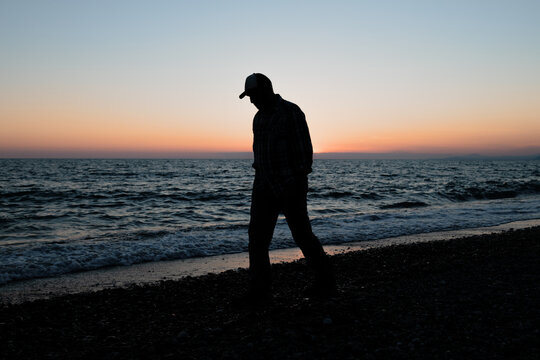 Silhouette Of A Man In A Cap Walking Along The Coastline