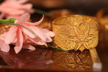 intricately handcrafted broad gold bangle with a pink lily flower near it.