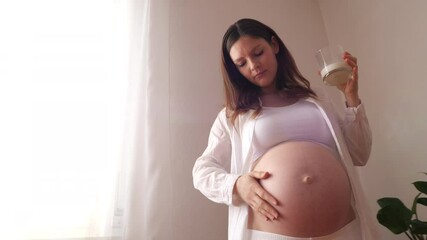 Pregnant woman holds a glass of milk in her hand while stroking her belly, camera movement moving away with a ray of light passing through the curtains of the house, slow motion video with copy space - Powered by Adobe
