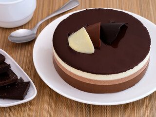 A Chocolate mousse cake on a white plate. Plate with pieces of chocolate, teaspoon and cup near it on bamboo napkin