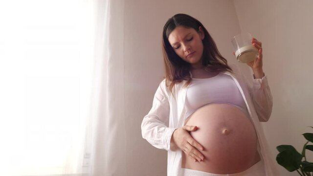 Pregnant woman holds a glass of milk in her hand while stroking her belly, camera movement moving away with a ray of light passing through the curtains of the house, slow motion video with copy space