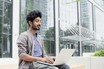 Handsome man using laptop in a city. Smiling indian male student working on computer outdoor. Modern lifestyle, connection, distance studying, communication online, student lifestyle, business concept