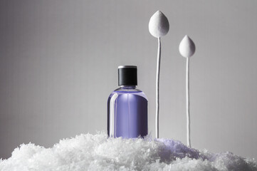 Glass spray bottle of purple perfume and decorative paper flower bud on white fluffy icy pile in studio on light grey background