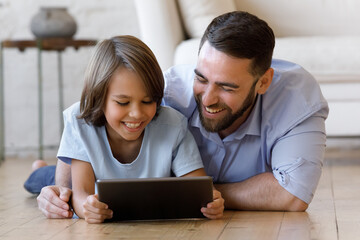 Obraz premium Happy millennial father and gen Z teenage son resting on heating floor at home, using tablet, watching, discussing funny movie, online series, talking on video call, laughing, shopping on Internet