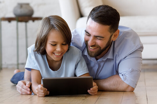 Happy Millennial Father And Gen Z Teenage Son Resting On Heating Floor At Home, Using Tablet, Watching, Discussing Funny Movie, Online Series, Talking On Video Call, Laughing, Shopping On Internet