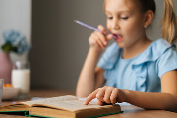 Close-up focus on foreground of thoughtful elementary child school girl doing homework and holding pen against mouth sitting at home table, selective focus. Homeschooling concept.