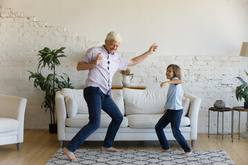 Happy active grandpa and cheerful grandkid dancing to music at home, having fun, spending active leisure time in living room, jumping together on carpeted floor, enjoying home party
