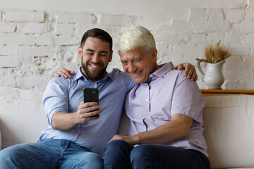 Cheerful excited grownup son and mature elder father taking selfie on smartphone, laughing,...