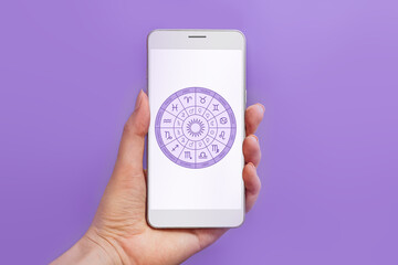 Woman's hand holding mobile phone with astrology application and zodiac circle on white screen isolated on lilac background